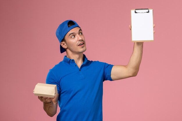 Vista frontal joven mensajero con capa uniforme azul sosteniendo un pequeño paquete de comida de entrega y un bloc de notas en la pared rosa claro