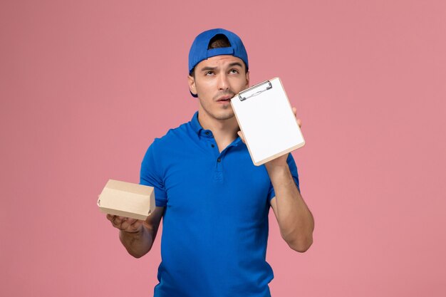 Vista frontal joven mensajero con capa uniforme azul sosteniendo un pequeño paquete de comida de entrega y un bloc de notas pensando en la pared rosa claro