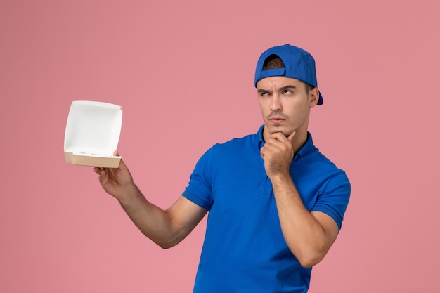Vista frontal joven mensajero con capa uniforme azul sosteniendo un pequeño paquete de comida de entrega vacía pensando en la pared rosa