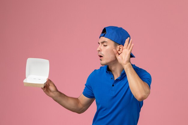 Vista frontal joven mensajero con capa uniforme azul sosteniendo un pequeño paquete de comida de entrega vacía y tratando de escuchar algo en la pared rosa