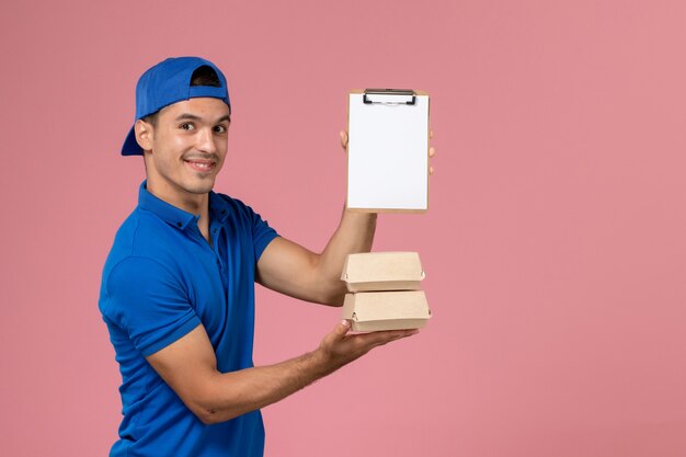 Vista frontal joven mensajero en capa uniforme azul sosteniendo pequeños paquetes de comida de entrega con bloc de notas en la pared rosa claro