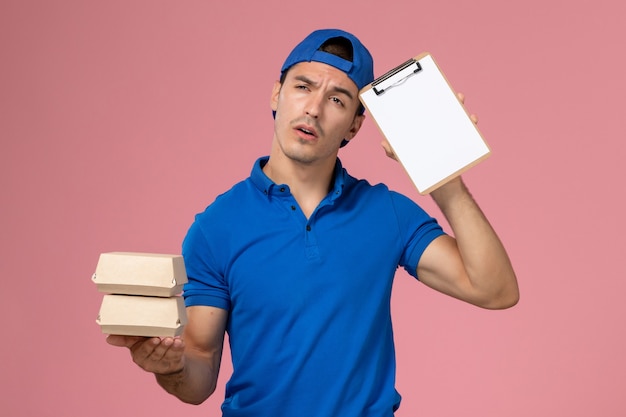 Vista frontal joven mensajero con capa uniforme azul sosteniendo pequeños paquetes de comida de entrega con bloc de notas pensando en la pared rosa claro