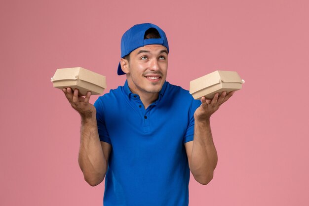 Vista frontal joven mensajero en capa uniforme azul sosteniendo pequeños paquetes de comida de entrega en la pared rosa
