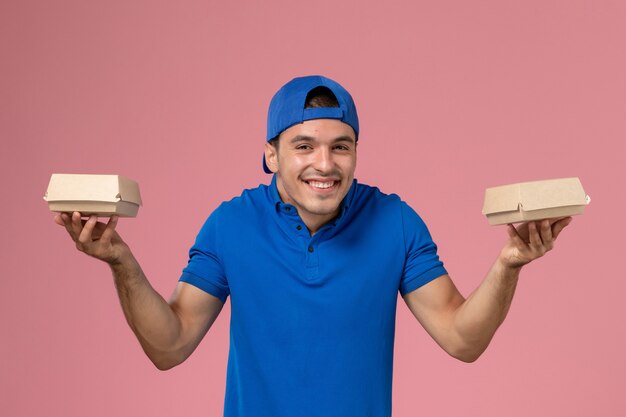 Foto gratuita vista frontal joven mensajero en capa uniforme azul sosteniendo pequeños paquetes de comida de entrega en la pared rosa