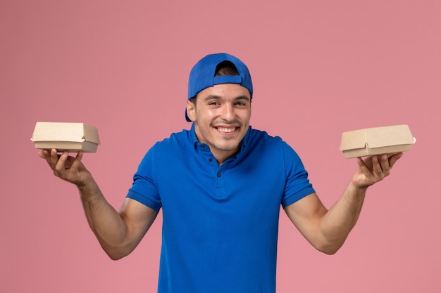 Vista frontal joven mensajero en capa uniforme azul sosteniendo pequeños paquetes de comida de entrega en la pared rosa