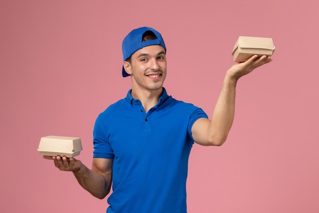 Vista frontal joven mensajero en capa uniforme azul sosteniendo pequeños paquetes de comida de entrega en la pared rosa