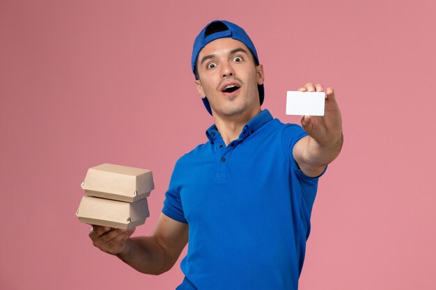 Vista frontal joven mensajero en capa uniforme azul sosteniendo pequeños paquetes de comida de entrega con tarjeta en la pared rosa claro