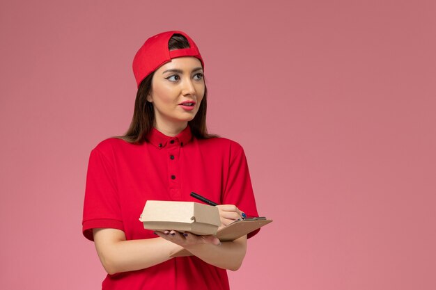 Vista frontal joven mensajero en capa uniforme roja con pequeño paquete de comida de entrega y bloc de notas en sus manos en la pared rosa claro