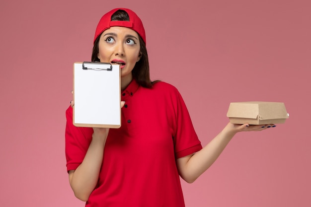 Vista frontal joven mensajero en capa uniforme roja con pequeño paquete de comida de entrega y bloc de notas en sus manos pensando en la pared rosa