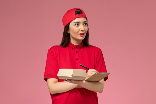 Vista frontal joven mensajero con capa de uniforme rojo con pequeño paquete de comida de entrega y bloc de notas con bolígrafo en sus manos en la pared rosa claro