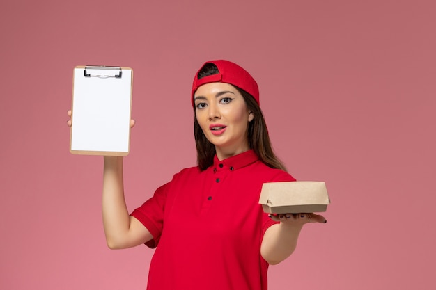 Vista frontal joven mensajero en capa uniforme rojo con pequeño paquete de comida de entrega y bloc de notas en sus manos en la pared rosa