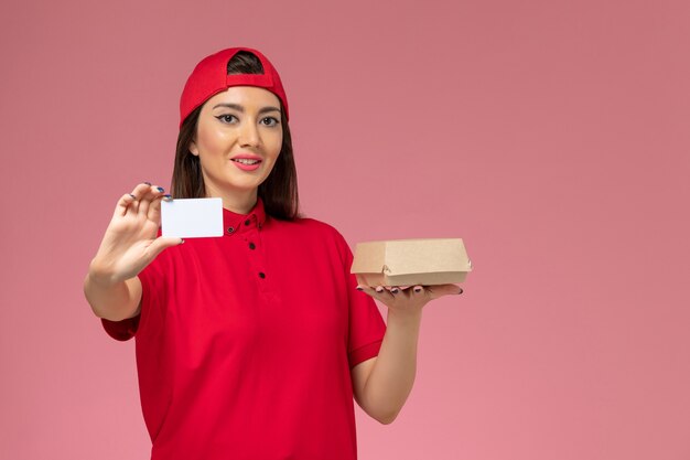 Vista frontal joven mensajero con capa de uniforme rojo con pequeño paquete de comida de entrega y tarjeta en sus manos en el empleado de entrega de servicio de escritorio rosa claro