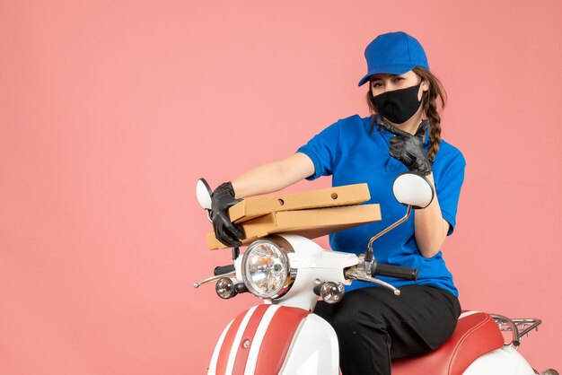 Vista frontal del joven mensajero confía en usar máscara médica y guantes sentado en scooter entregando pedidos sobre fondo de melocotón pastel