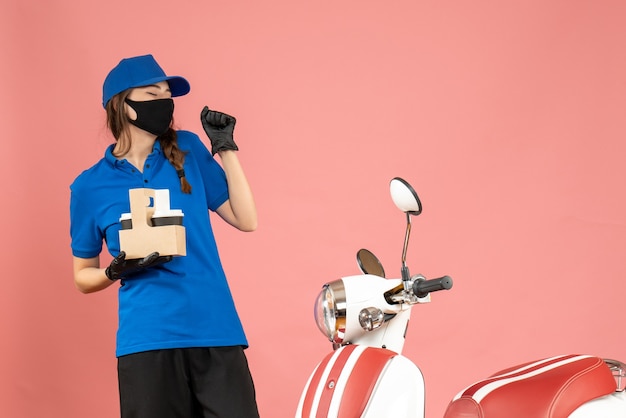 Vista frontal de la joven mensajero feliz con guantes de máscara médica de pie junto a la motocicleta sosteniendo pequeños pasteles de café sobre fondo de color melocotón pastel