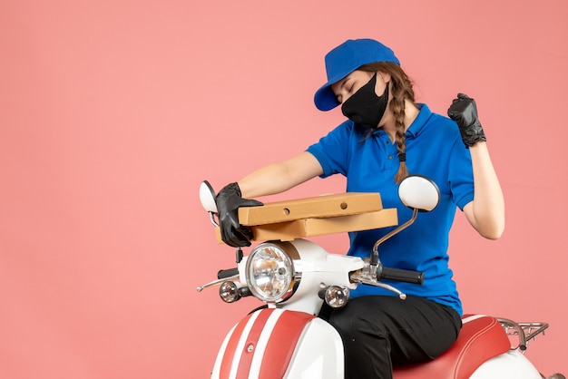 Vista frontal del joven mensajero femenino emocional feliz con máscara médica y guantes sentado en scooter entregando pedidos sobre fondo melocotón pastel