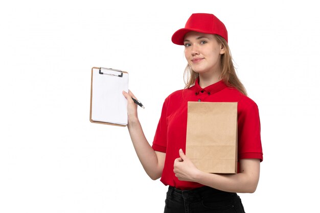 Una vista frontal joven mensajero femenino trabajadora del servicio de entrega de alimentos sonriendo sosteniendo el bloc de notas y paquete de alimentos en blanco