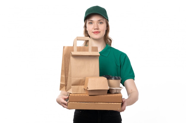 Una vista frontal joven mensajero femenino en uniforme verde con paquetes de alimentos de taza de café y sonriendo en blanco