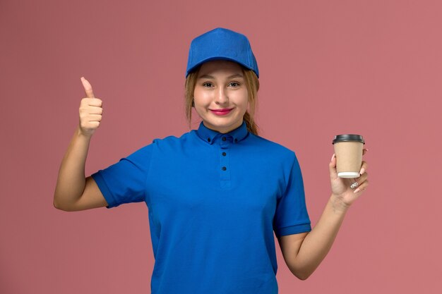 Vista frontal joven mensajero sonriente en uniforme azul posando sosteniendo una taza de café de entrega marrón, mujer de entrega uniforme de trabajo de servicio