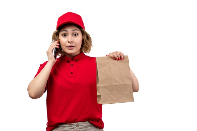 Una vista frontal joven mensajero trabajadora del servicio de entrega de alimentos con paquete de alimentos y hablando por teléfono en blanco