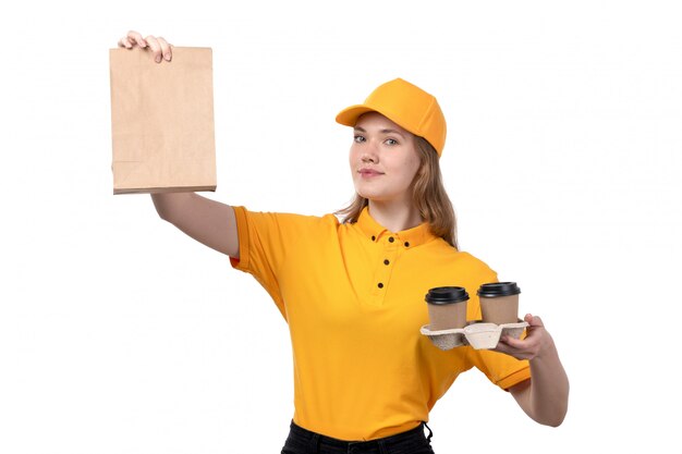 Una vista frontal joven mensajero trabajadora del servicio de entrega de alimentos sonriendo sosteniendo paquetes de alimentos y tazas de café en blanco