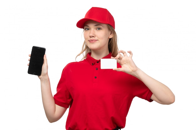 Una vista frontal joven mensajero trabajadora del servicio de entrega de alimentos sonriendo sosteniendo la tarjeta whtie y teléfono inteligente en blanco