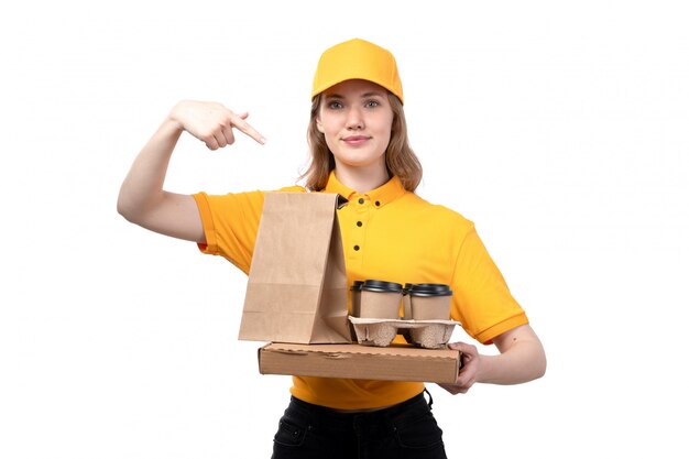 Una vista frontal joven mensajero trabajadora del servicio de entrega de alimentos sosteniendo tazas de café paquetes de alimentos y sonriendo en blanco