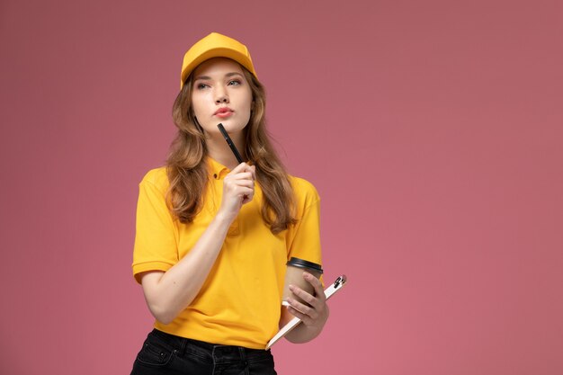 Vista frontal joven mensajero en uniforme amarillo con bloc de notas y café en el escritorio de color rosa oscuro uniforme servicio de entrega trabajadora