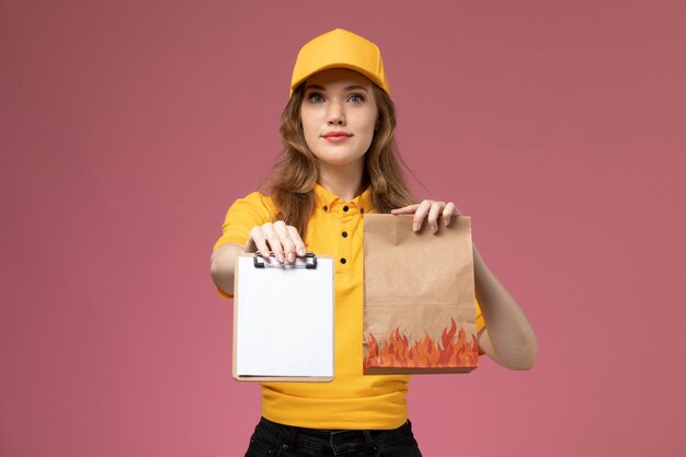 Vista frontal joven mensajero en uniforme amarillo con bloc de notas de paquete de comida de entrega en el servicio de entrega uniforme de trabajo de fondo rosa