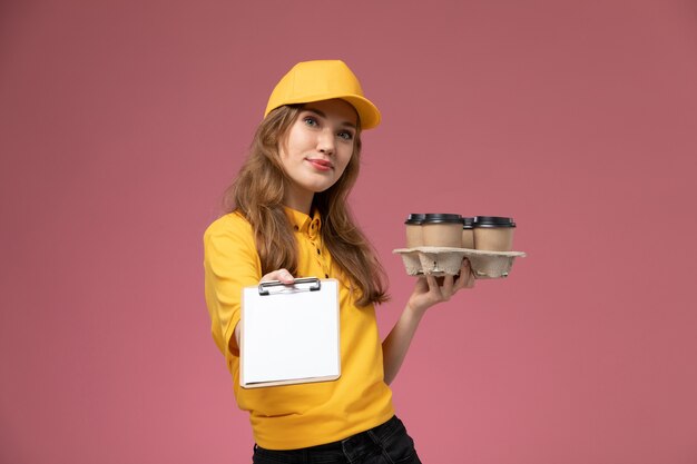 Vista frontal joven mensajero en uniforme amarillo con café de entrega y bloc de notas con una sonrisa en el trabajo de escritorio rosa trabajador de servicio de entrega uniforme