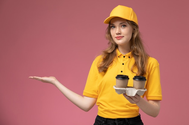 Vista frontal joven mensajero en uniforme amarillo con café de entrega posando en el trabajo de escritorio rosa trabajador de servicio de entrega uniforme