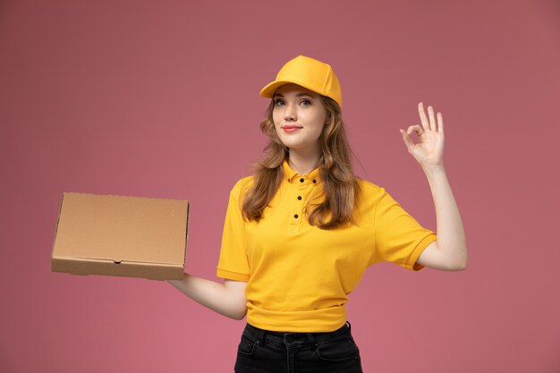 Vista frontal joven mensajero en uniforme amarillo con caja de comida de entrega con sonrisa en el trabajo de escritorio rosa trabajador de servicio de entrega uniforme
