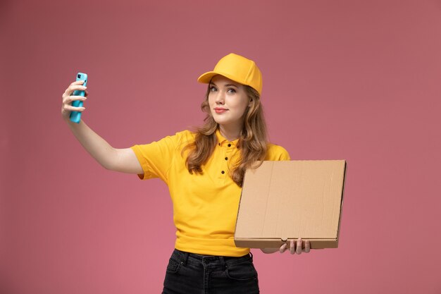 Vista frontal joven mensajero en uniforme amarillo con caja de comida tomando una foto con él en el escritorio de color rosa oscuro trabajador de servicio de entrega uniforme de trabajo