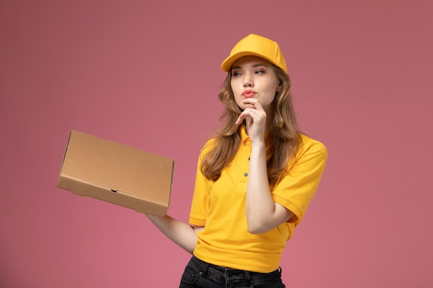 Vista frontal joven mensajero en uniforme amarillo capa amarilla con caja de entrega de alimentos en el color de servicio de trabajo de entrega uniforme de fondo rosa oscuro