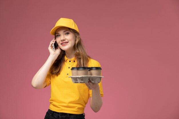 Vista frontal joven mensajero en uniforme amarillo capa amarilla hablando por teléfono en el color de servicio de trabajo de entrega uniforme de fondo rosa oscuro