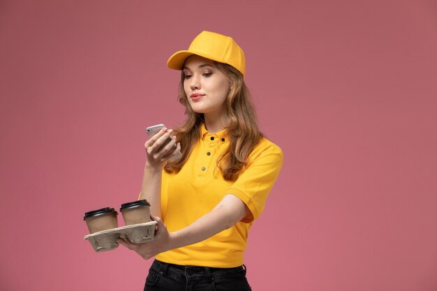 Vista frontal joven mensajero en uniforme amarillo capa amarilla sosteniendo tazas de café usando su teléfono en el escritorio de color rosa oscuro servicio de entrega uniforme trabajadora