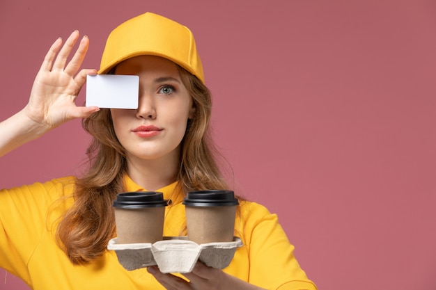 Vista frontal joven mensajero en uniforme amarillo con entrega de café y tarjeta blanca en el trabajo de escritorio rosa trabajador de servicio de entrega uniforme