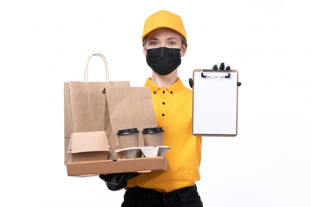 Una vista frontal joven mensajero en uniforme amarillo guantes negros y máscara negra sosteniendo cajas de pizza y entrega de bloc de notas