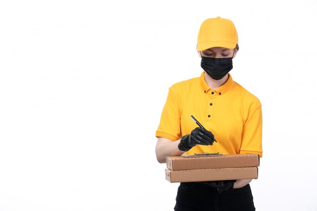 Una vista frontal joven mensajero en uniforme amarillo guantes negros y máscara negra sosteniendo paquetes de alimentos orden de escritura