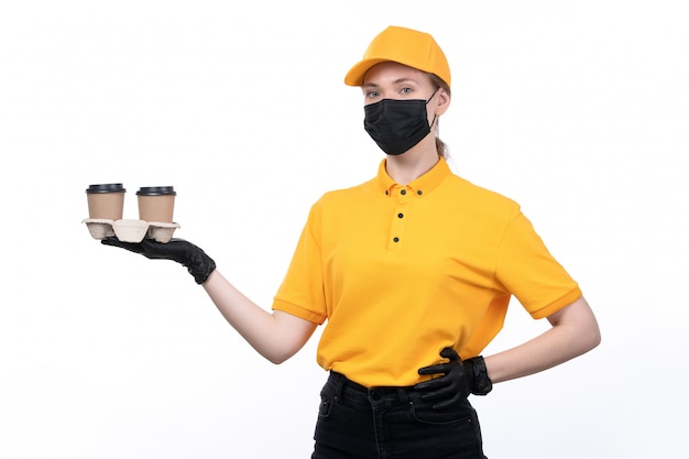 Una vista frontal joven mensajero en uniforme amarillo guantes negros y máscara negra sosteniendo tazas de café entregando