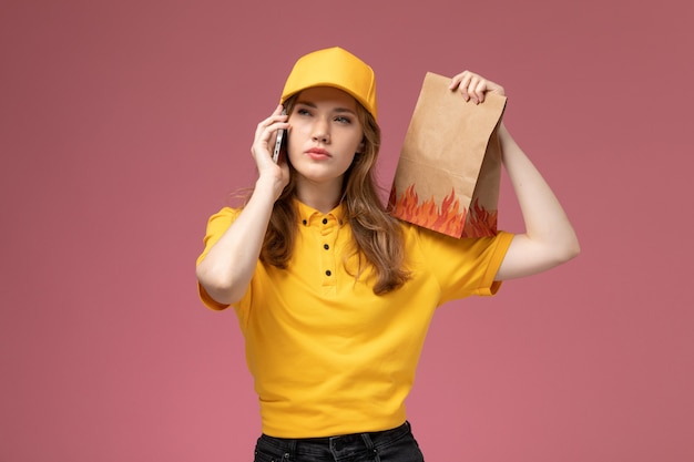 Vista frontal joven mensajero en uniforme amarillo con paquete de comida de entrega y hablando por teléfono en el trabajo de escritorio rosa trabajador de servicio de entrega uniforme