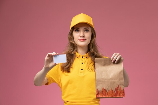 Vista frontal joven mensajero en uniforme amarillo con paquete de comida de entrega y tarjeta de plástico blanco en el trabajo de escritorio rosa trabajador de servicio de entrega uniforme