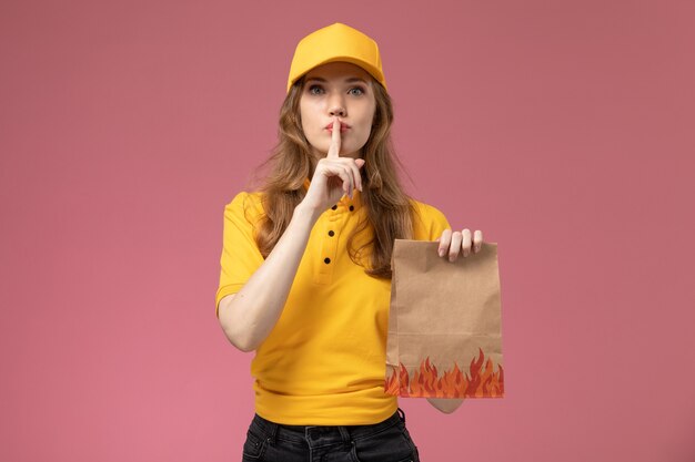 Vista frontal joven mensajero en uniforme amarillo con paquete de entrega de alimentos en el escritorio de color rosa oscuro servicio de entrega uniforme trabajadora