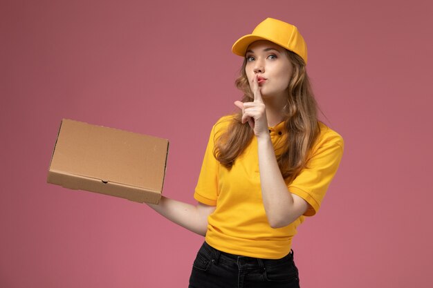 Vista frontal joven mensajero en uniforme amarillo sosteniendo el paquete de comida de entrega y mostrando signo de silencio en el trabajo de escritorio rosa trabajador de servicio de entrega uniforme