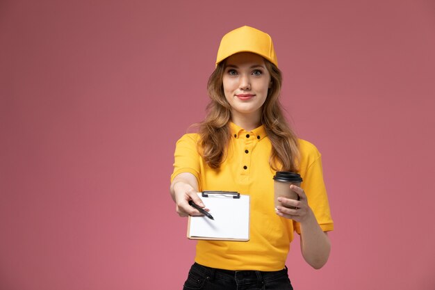 Vista frontal joven mensajero en uniforme amarillo sosteniendo una taza de café de plástico con el bloc de notas en el escritorio rosa oscuro trabajador de servicio de trabajo de entrega uniforme
