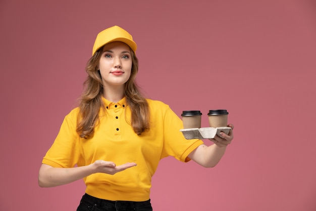 Vista frontal joven mensajero en uniforme amarillo sosteniendo tazas de café de entrega en el fondo rosa trabajo de escritorio uniforme trabajador de servicio de entrega