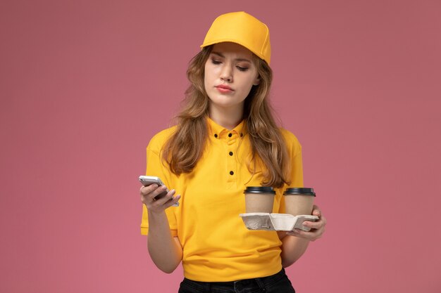 Vista frontal joven mensajero en uniforme amarillo sosteniendo tazas de café mientras usa su teléfono sobre fondo rosa trabajo de escritorio uniforme trabajador de servicio de entrega