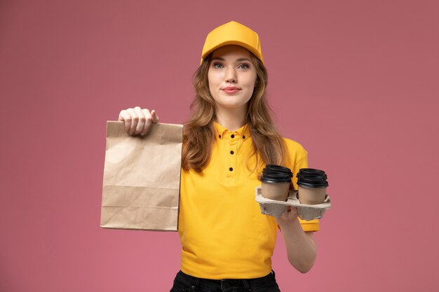 Vista frontal joven mensajero en uniforme amarillo sosteniendo tazas de café y paquete de comida en el escritorio de color rosa oscuro uniforme trabajador de servicio de trabajo de entrega