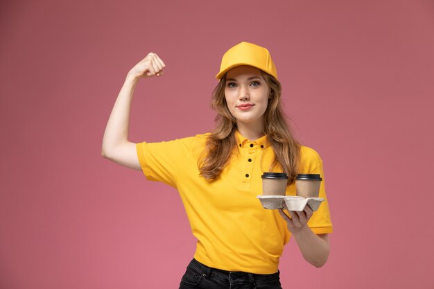 Vista frontal joven mensajero en uniforme amarillo sosteniendo tazas de café de plástico marrón y flexionando en el trabajo de escritorio de color rosa oscuro uniforme entrega trabajador de servicio de color
