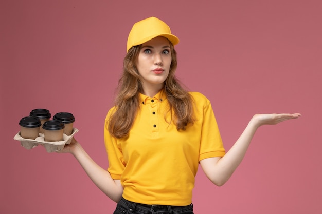 Vista frontal joven mensajero en uniforme amarillo sosteniendo tazas de café de plástico marrón en el fondo rosa trabajo uniforme entrega color trabajador de servicio
