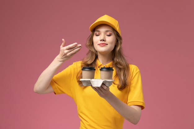 Vista frontal joven mensajero en uniforme amarillo sosteniendo tazas de café de plástico marrón oliéndolas sobre fondo rosa trabajo uniforme entrega color trabajador de servicio